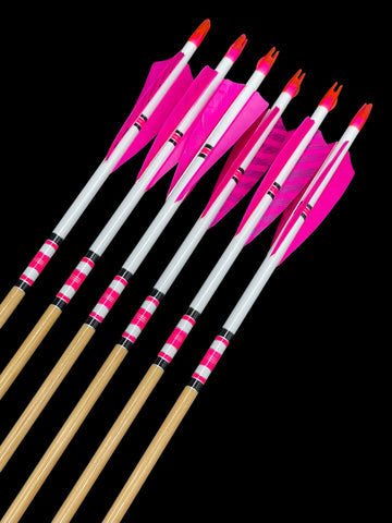 A collection of seven archery arrows with wooden shafts, white and pink fletching, and pink nocks and tips, arranged diagonally against a solid black background. The arrows feature a distinctive pink and white striped pattern near the fletching.