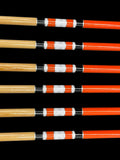 A set of six wooden arrows with orange shafts, featuring black, white, and orange bands near the fletching area, neatly aligned against a black background. The natural wood grain is visible at the ends of the arrows.
